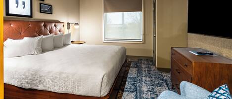 A bedroom with a large bed, a wooden dresser, a television, and a window with blinds.