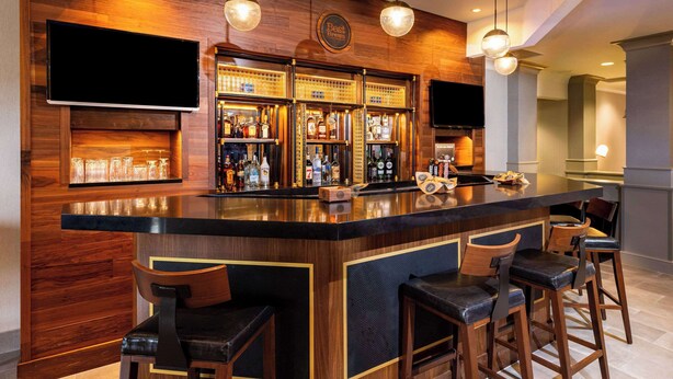 A bar area with a wooden bar counter, multiple stools, and shelves stocked with bottles. - Radisson Hotel Fairview Heights - St. Louis (Fairview Heights)