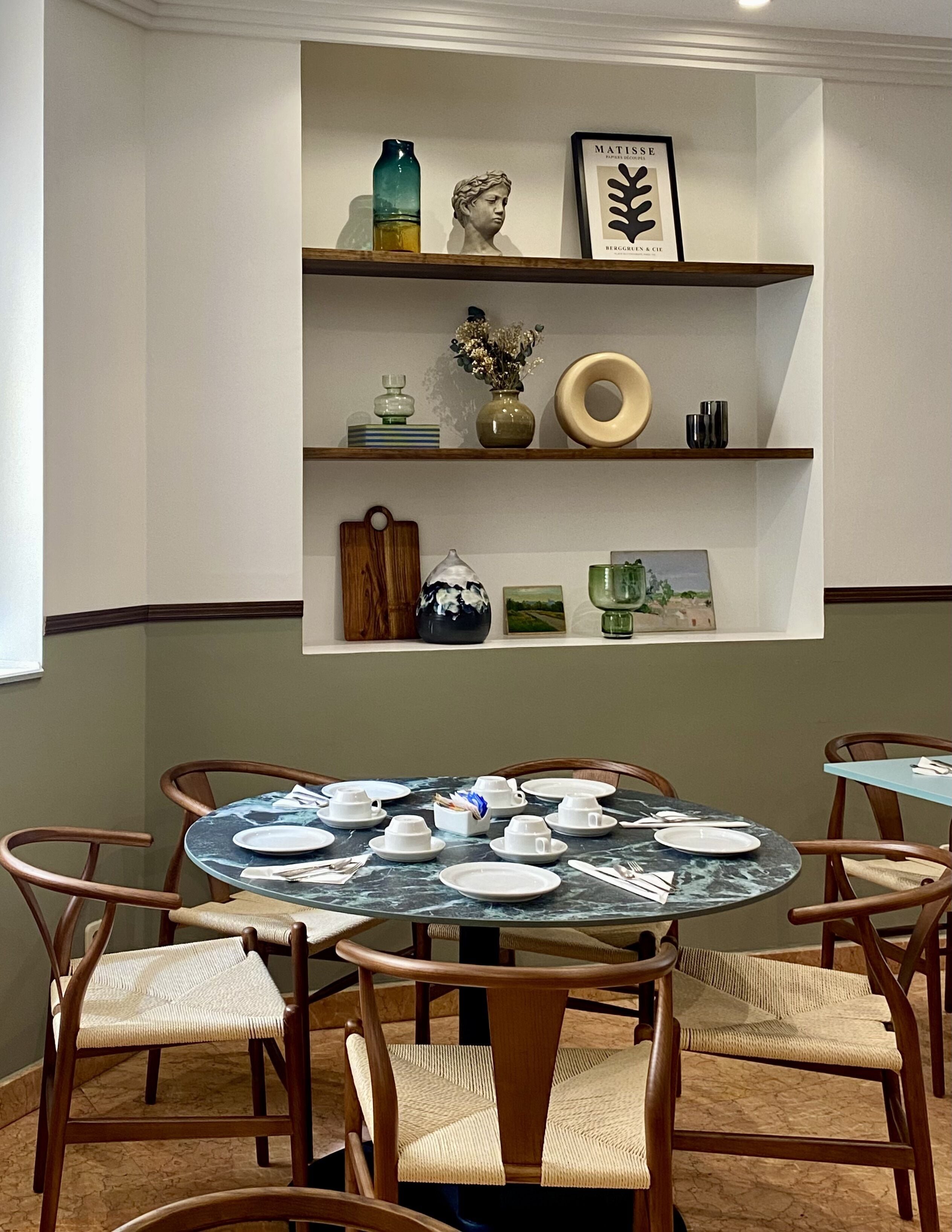 Un comedor con una mesa redonda dispuesta para seis comensales, rodeada por seis sillas de mimbre. Sobre la mesa, hay dos estanterías con objetos decorativos como un jarrón, un busto y una obra de arte enmarcada.