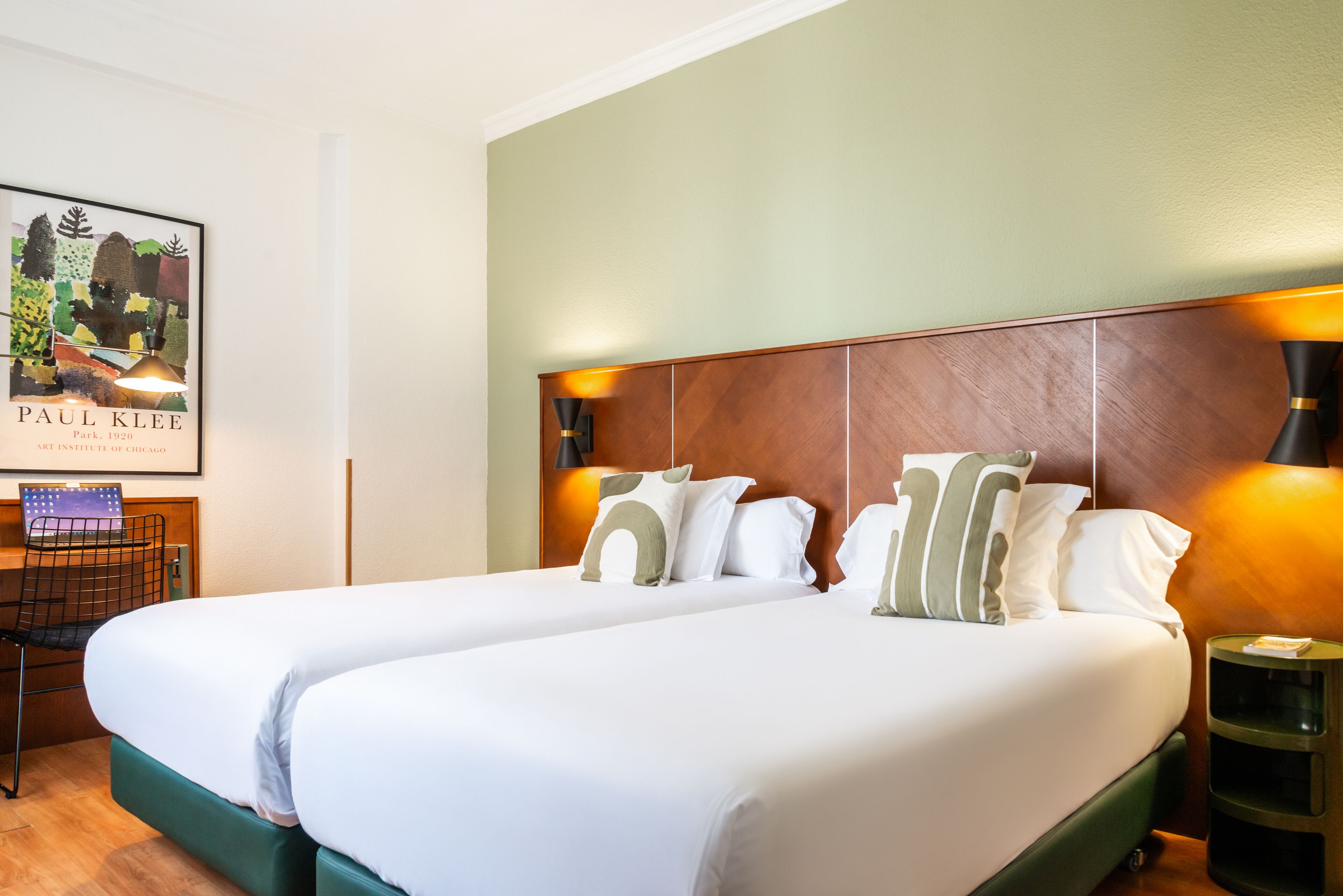 Habitación de hotel con dos camas, un escritorio con una computadora portátil, una obra de arte enmarcada y una mesita de noche con una lámpara.