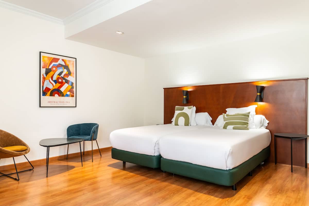 A modern hotel room with a large bed, two bedside tables, a chair, and a wooden headboard.