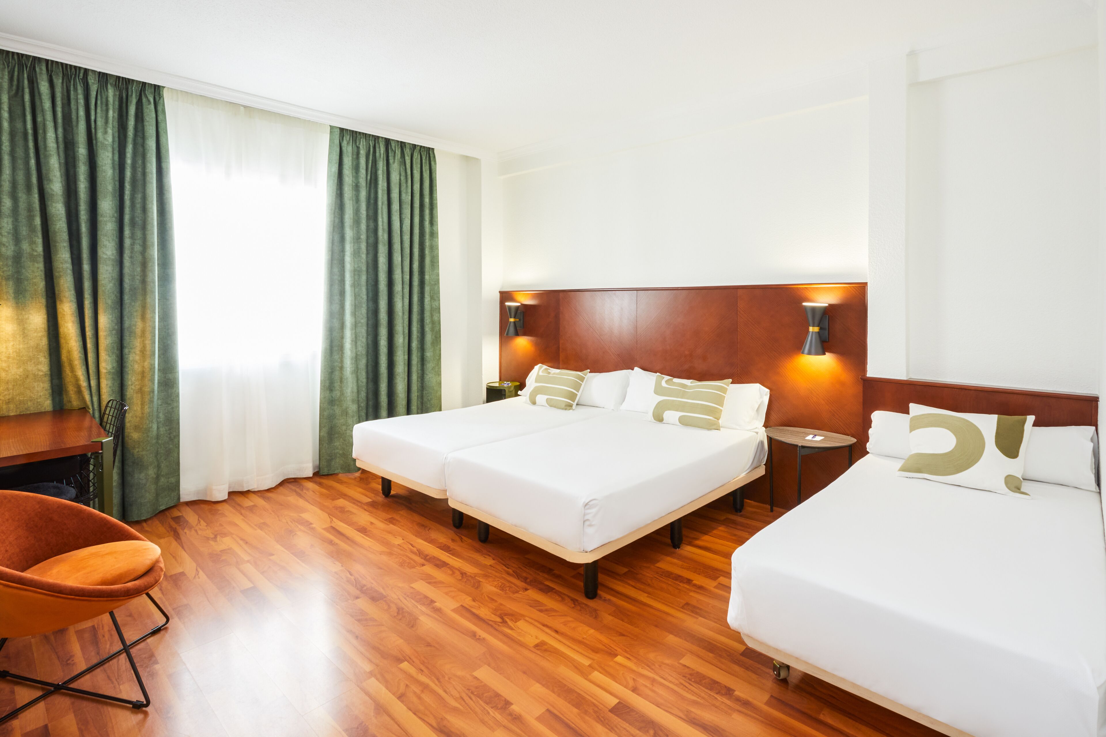 Habitació d'hotel amb dos llits, un sòl de fusta, una taula de treball, una cadira i una finestra amb cortines.