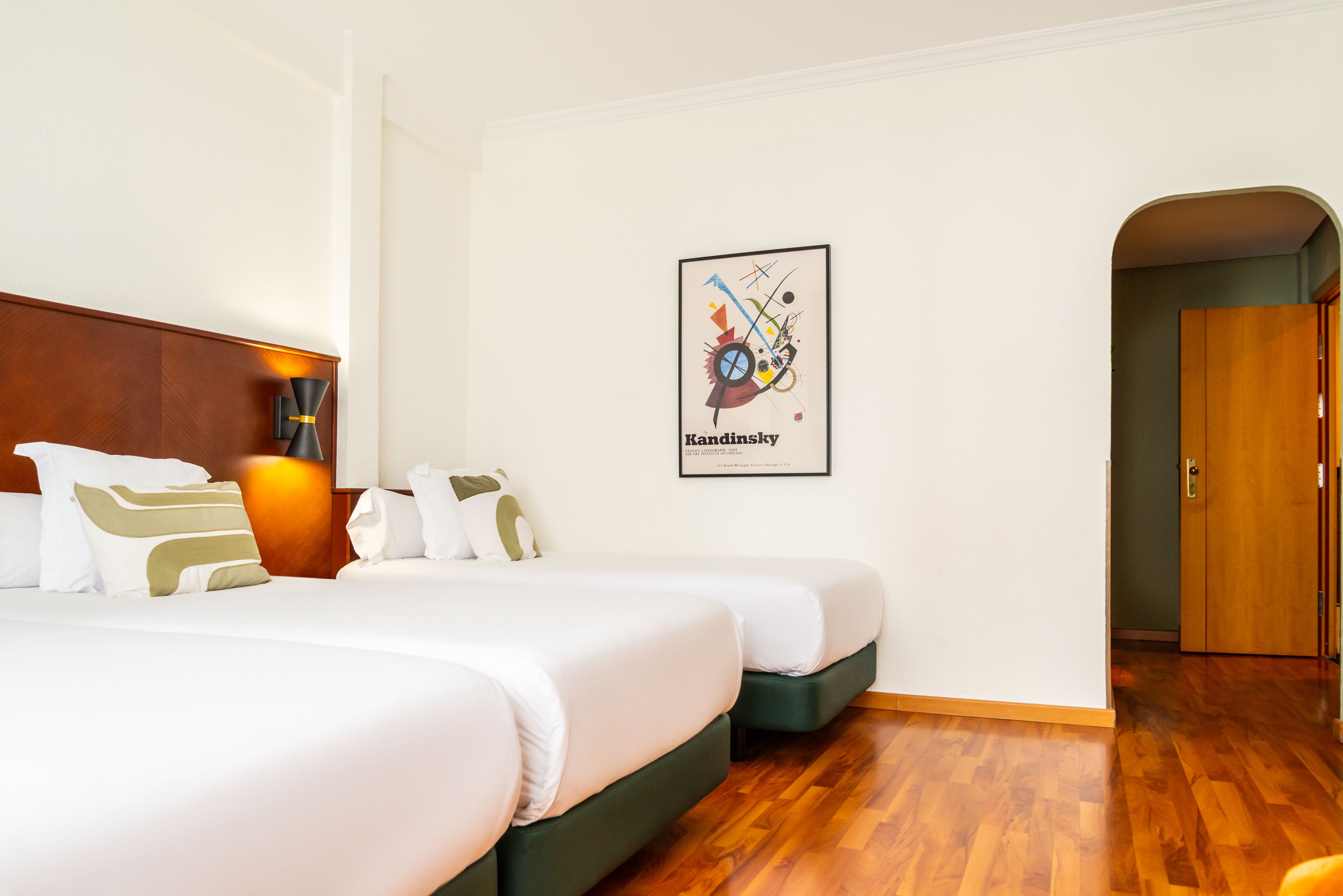 A hotel room with two beds, a wooden floor, a framed artwork on the wall, and a door leading to another room.