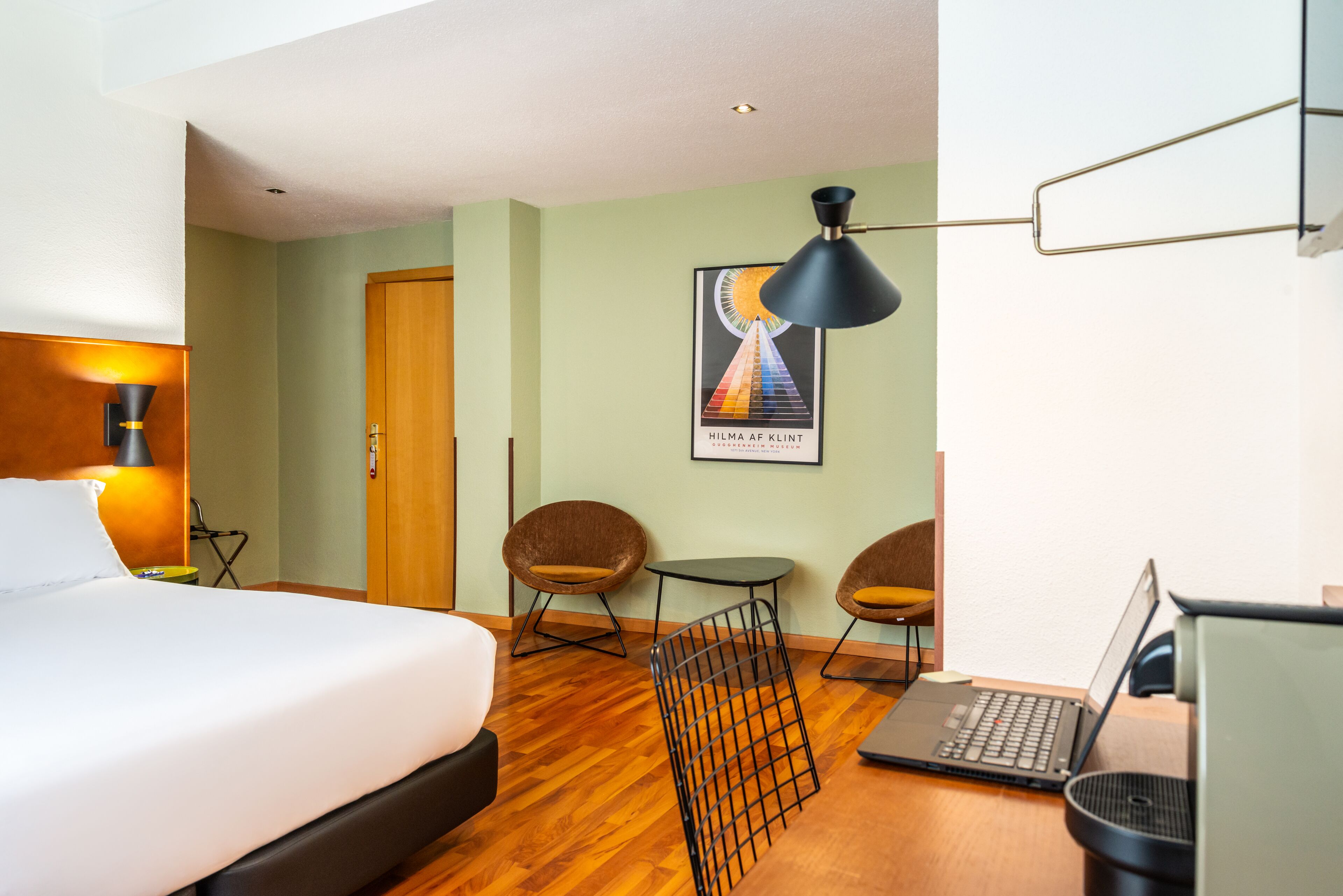 Habitación de hotel con una cama, un escritorio con una computadora portátil, dos sillas, una mesita y un cartel en la pared.