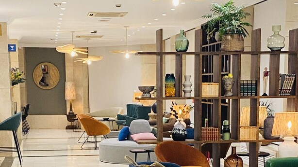 A modern interior with a central seating area featuring blue and orange chairs, a green sofa, and a wicker chair. There are ceiling fans, a plant, and a bookshelf with decorative items. - Hôtel Casón del Tormes by HappyCulture (Madrid)