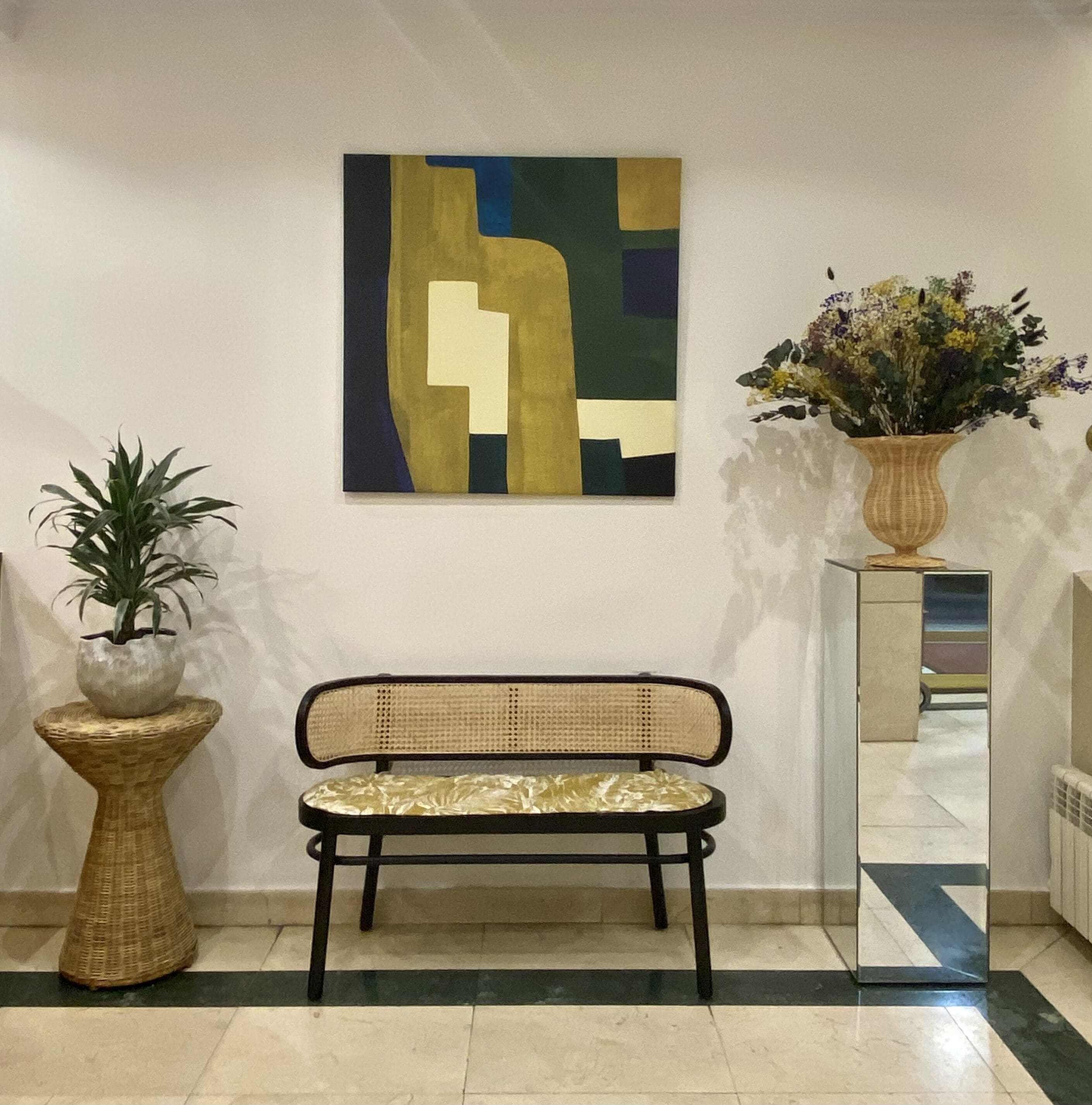 Un banco con respaldo tejido y asiento acolchado, una planta en maceta y un cuadro abstracto colgado en la pared.