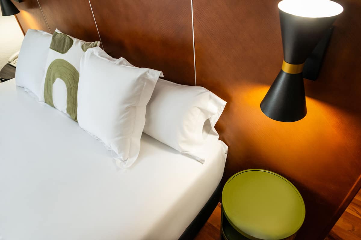 A neatly made bed with white linens and green and white pillows, a green bedside table, and a wall-mounted lamp.