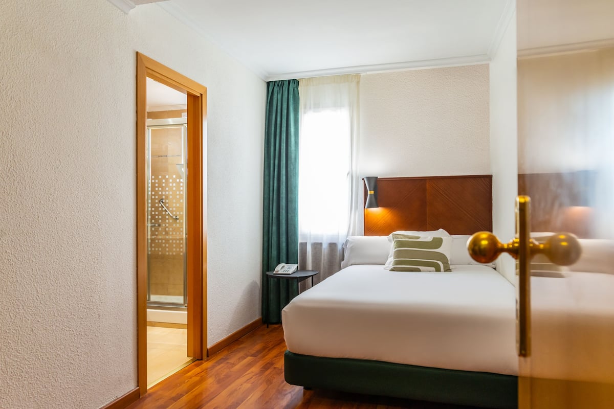 A hotel room with a wooden floor, a bed with white and green bedding, a nightstand with a lamp, and a window with curtains.