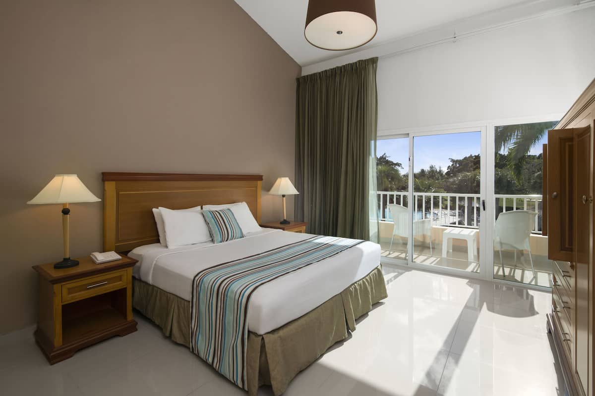 Hotelkamer met een groot bed, twee bedlampen, een houten nachtkastje en een glazen balkon met uitzicht op palmbomen.