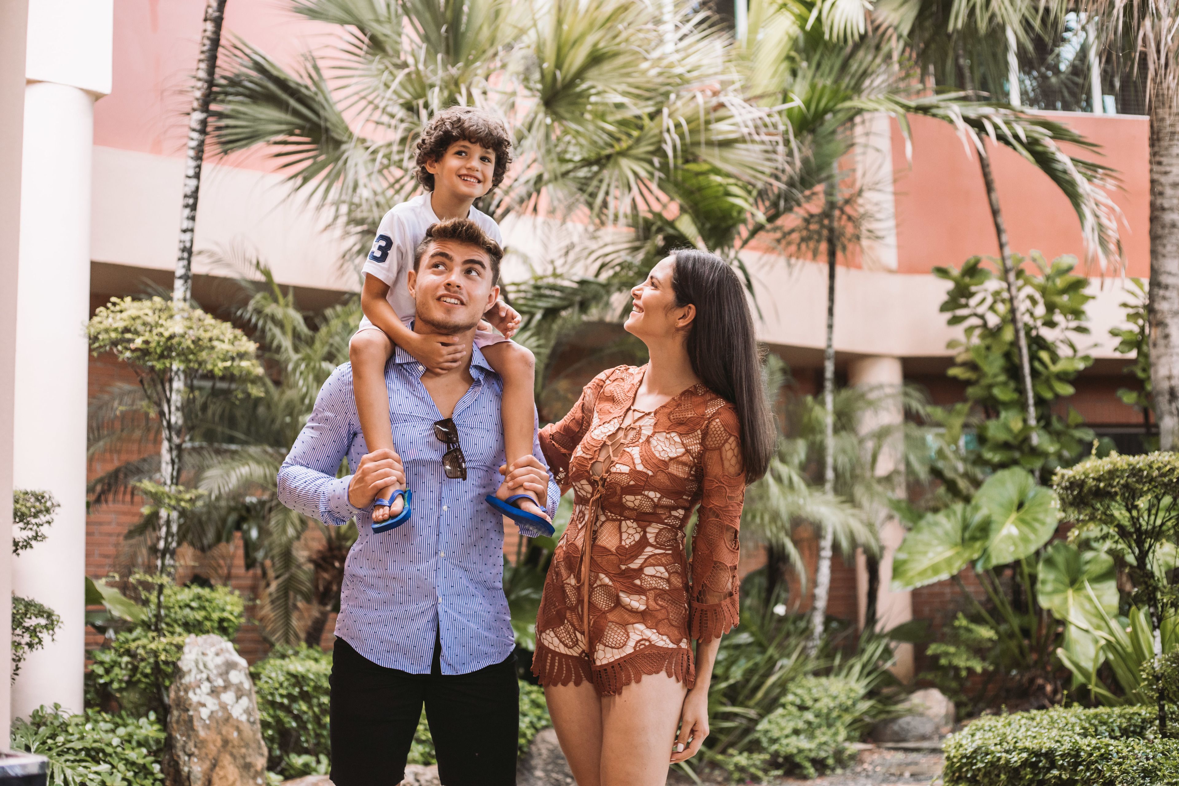 Una famiglia con un bambino sulle spalle, in un cortile con palme e un edificio sullo sfondo.