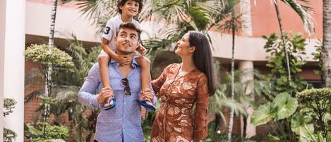 A family with a child on an adult's shoulders, standing in a courtyard with palm trees and a building in the background.