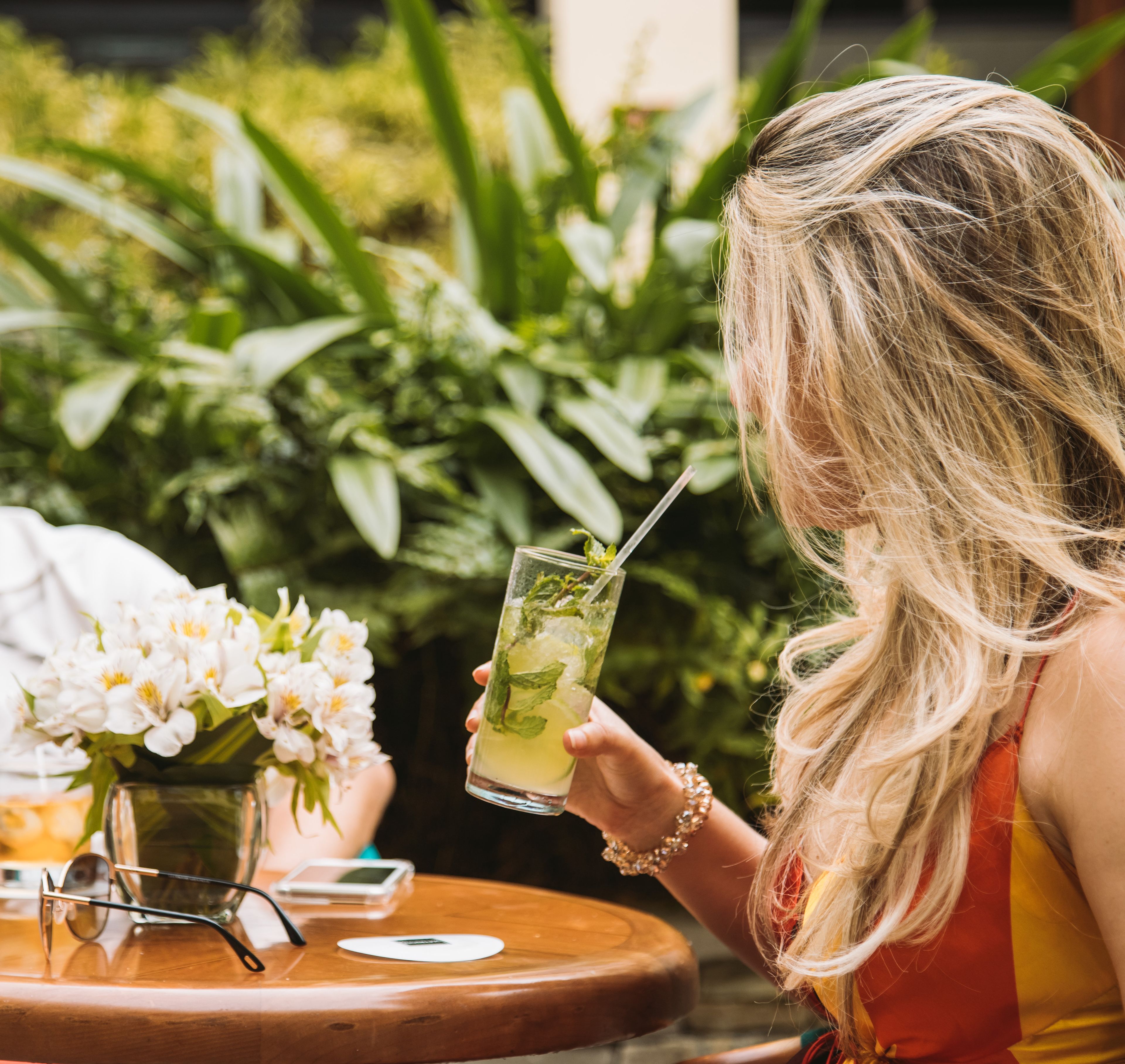 A person holding a glass with a straw, seated at a wooden table with a vase of flowers and a smartphone.