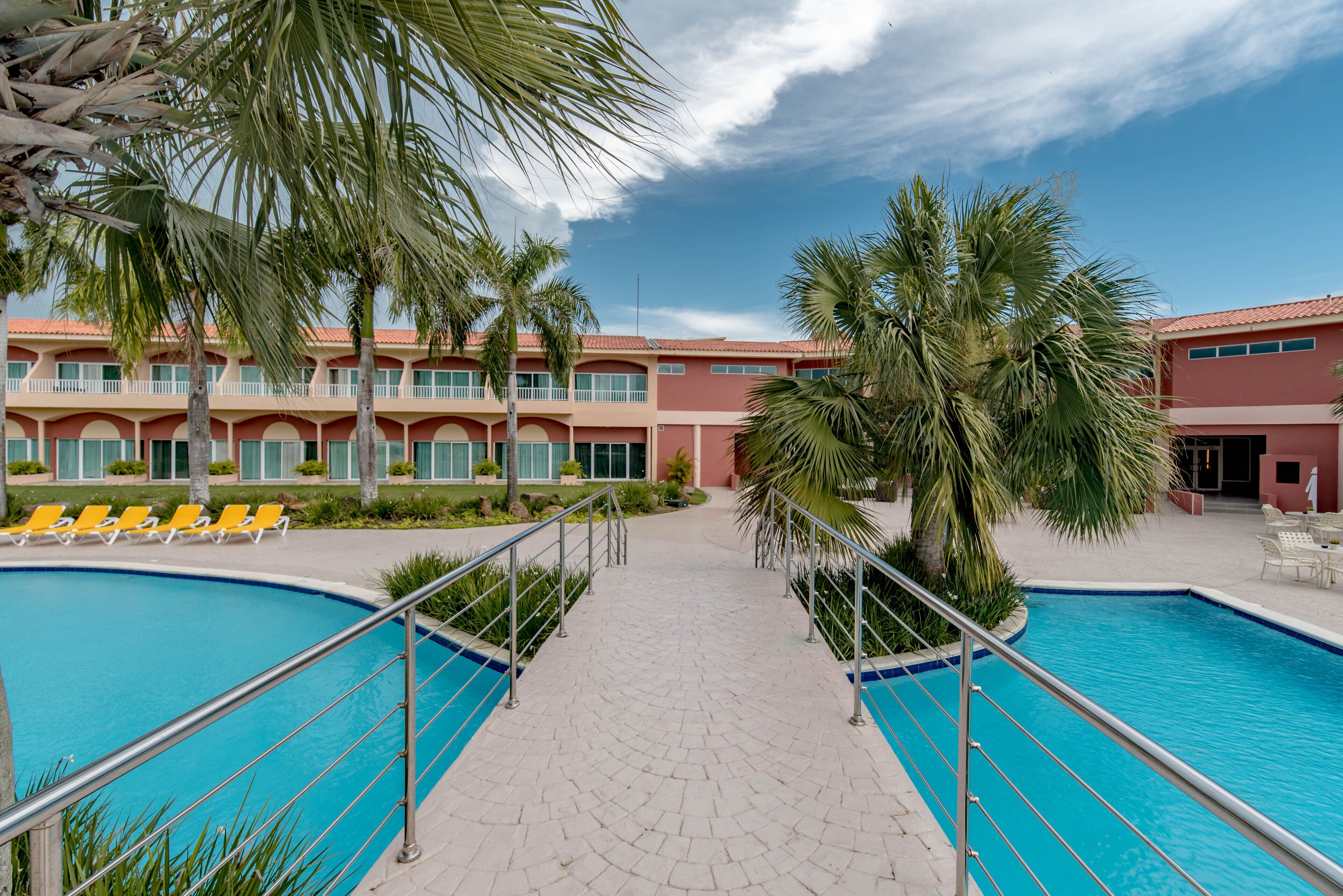 A swimming pool with a tiled walkway, palm trees, and a building with balconies in the background.