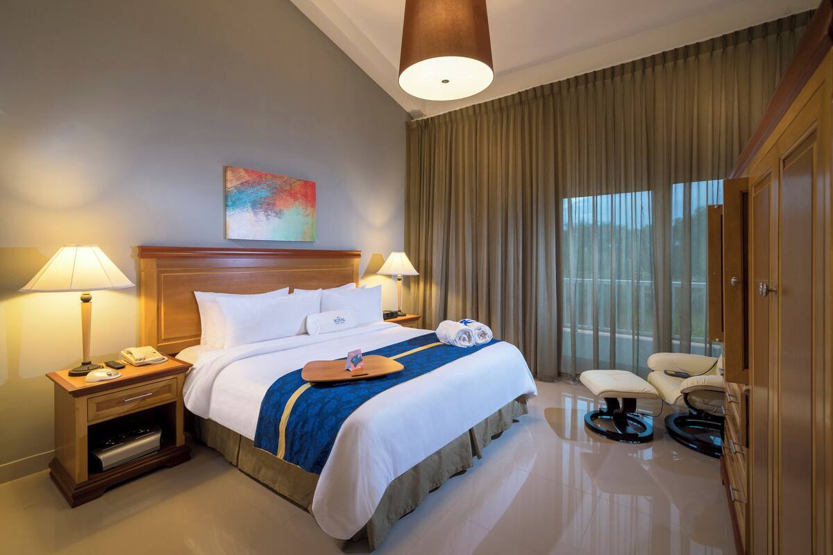 Een hotelkamer met een groot bed, twee nachtkastjes met lampen, een stoel, een bureau en een groot raam met gordijnen.