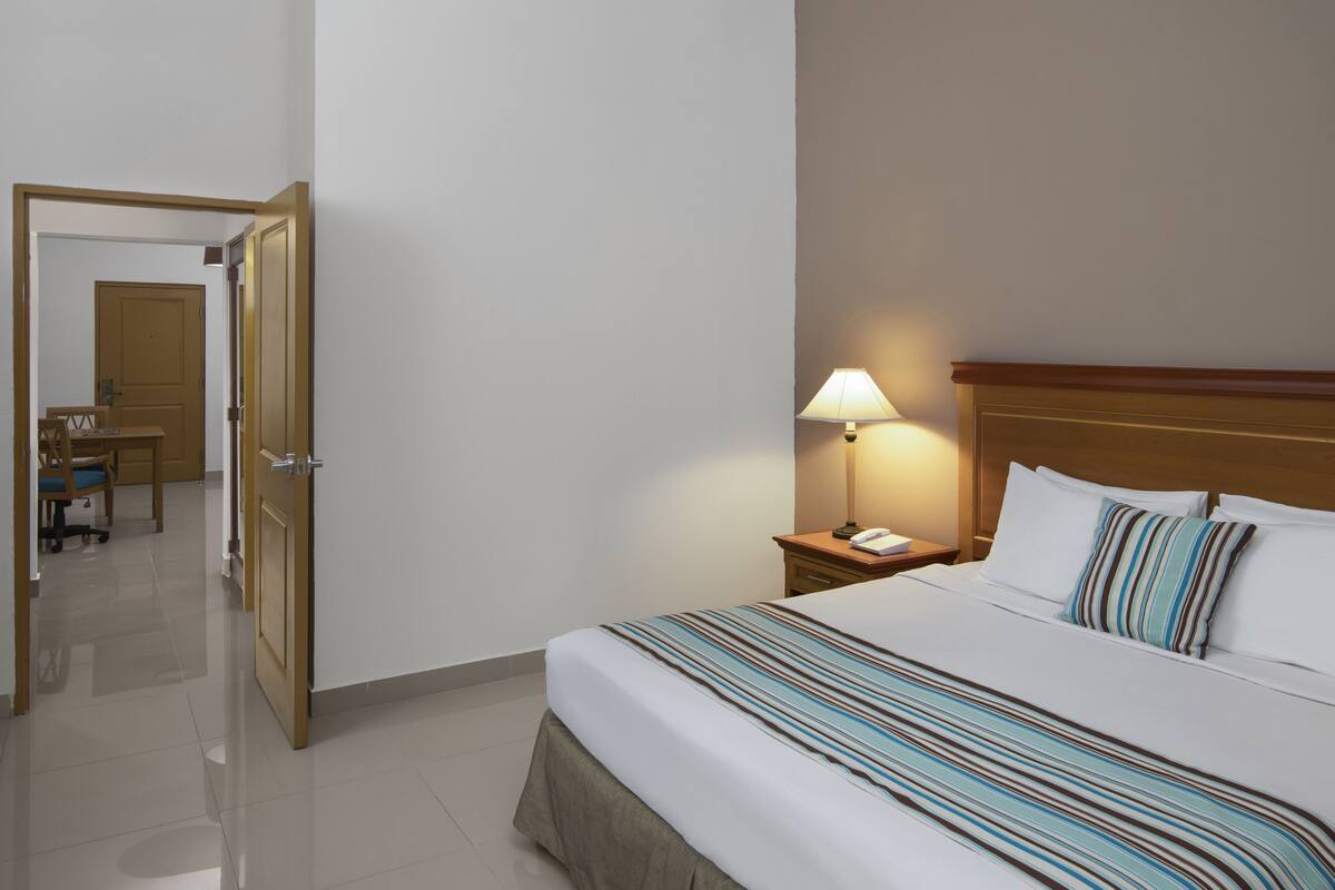 Een hotelkamer met een bed, nachtkastje, lamp en een deur naar een bureau.