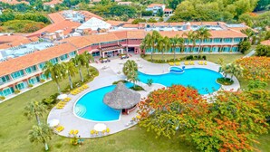 Vue aérienne d’un complexe hôtelier doté d’une grande piscine centrale, de plusieurs bâtiments et d’un environnement verdoyant.