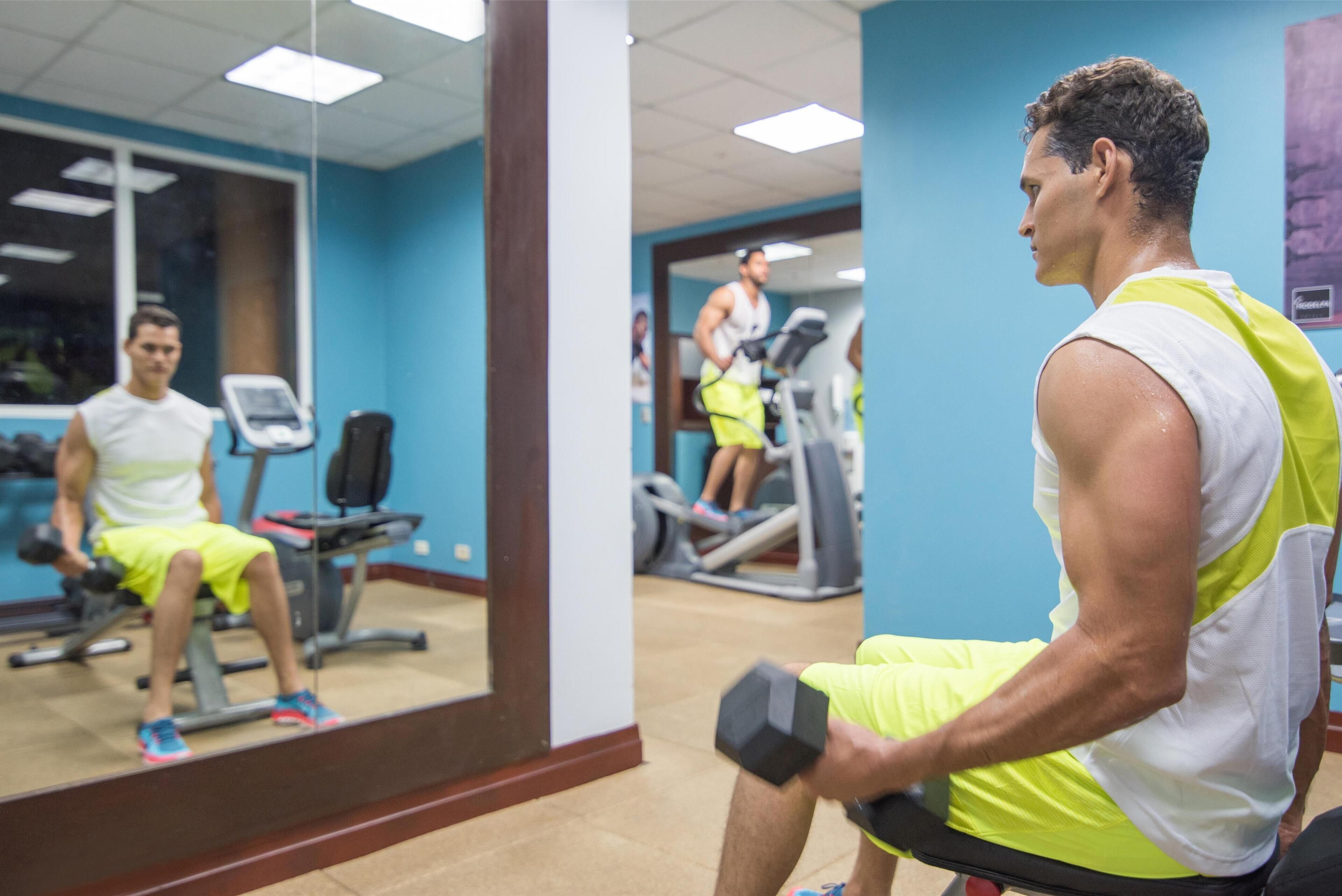 Ein Fitnessraum mit Trainingsgeräten, einem Spiegel und einer Person, die mit Kurzhanteln trainiert.