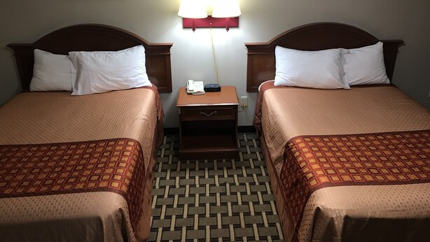 A hotel room with two beds, a nightstand with a lamp, and a patterned floor. - AmeriVu Inn & Suites (Wolcott)