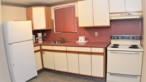 A compact kitchen with a refrigerator, sink, stove, and wooden cabinets. - AmeriVu Inn & Suites (Wolcott)