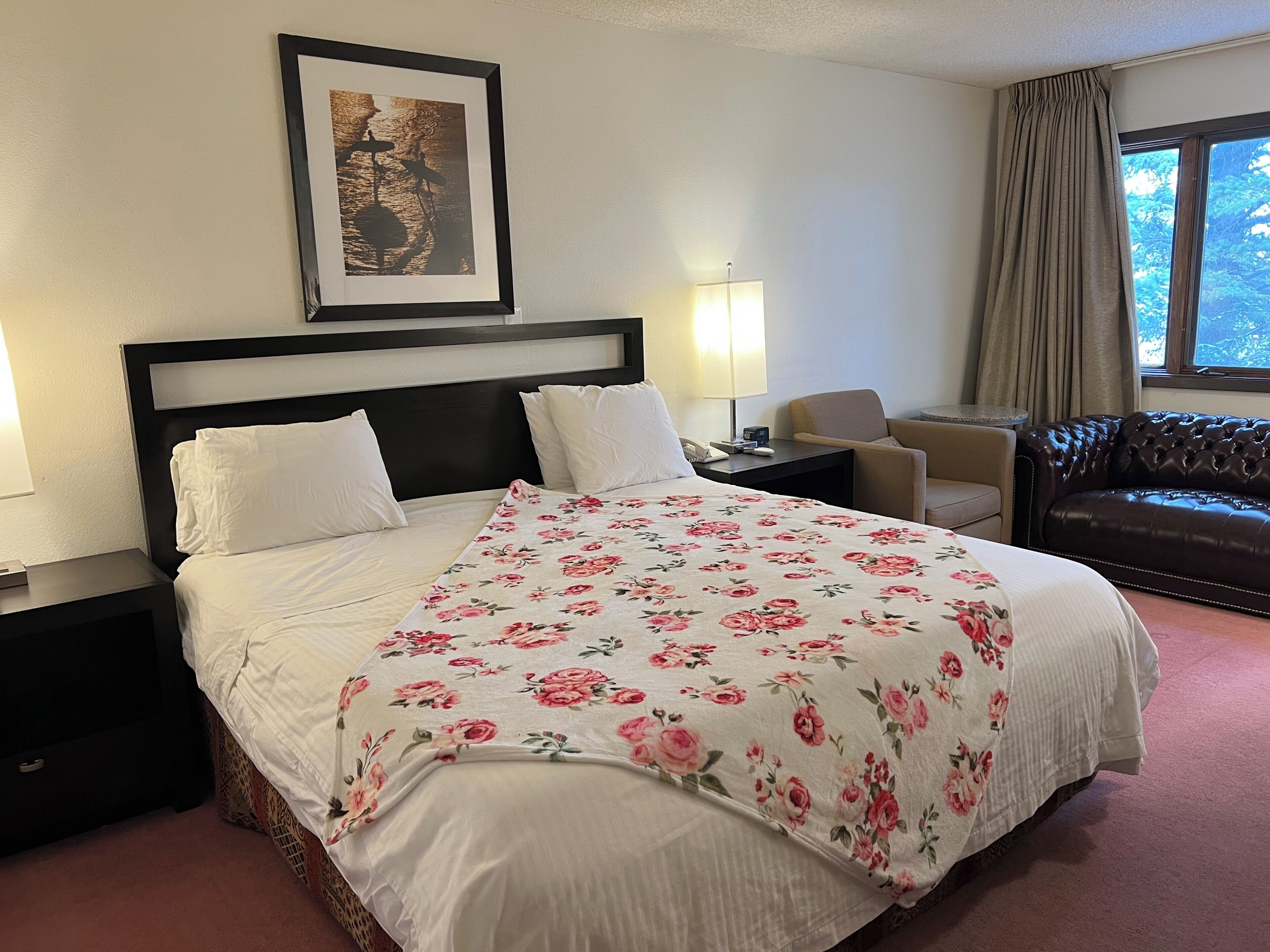A hotel room with a bed, a nightstand, a chair, a sofa, a lamp, and a window with curtains.