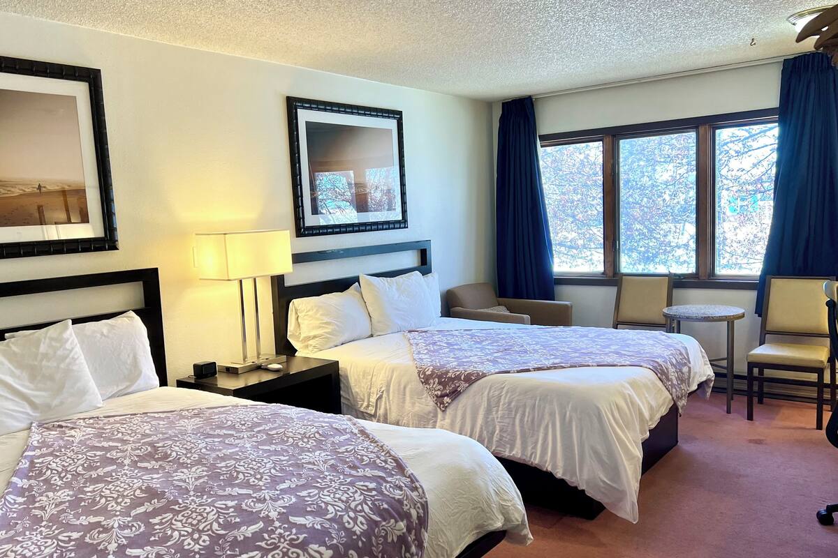 A hotel room with two beds, a desk, a chair, a small table, and a window with curtains.