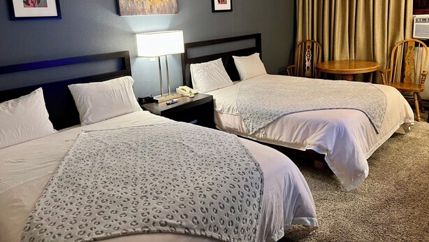 A hotel room with two beds, a nightstand with a lamp, a chair, and two framed artworks on the wall. - Americas Best Value Inn Laramie (Laramie)