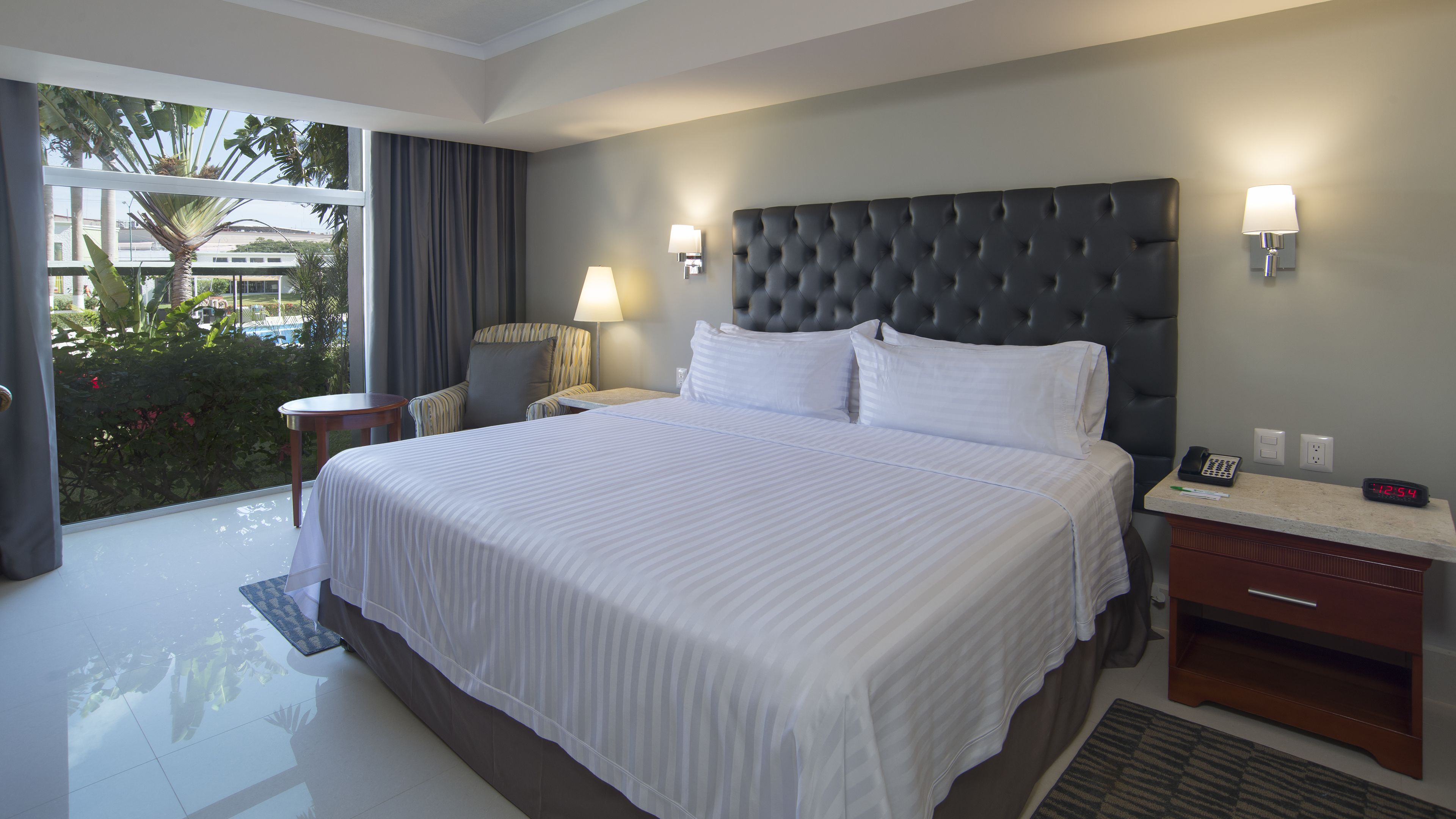 A hotel room with a large bed, two bedside tables, a chair, a lamp, and a view of the outdoors.