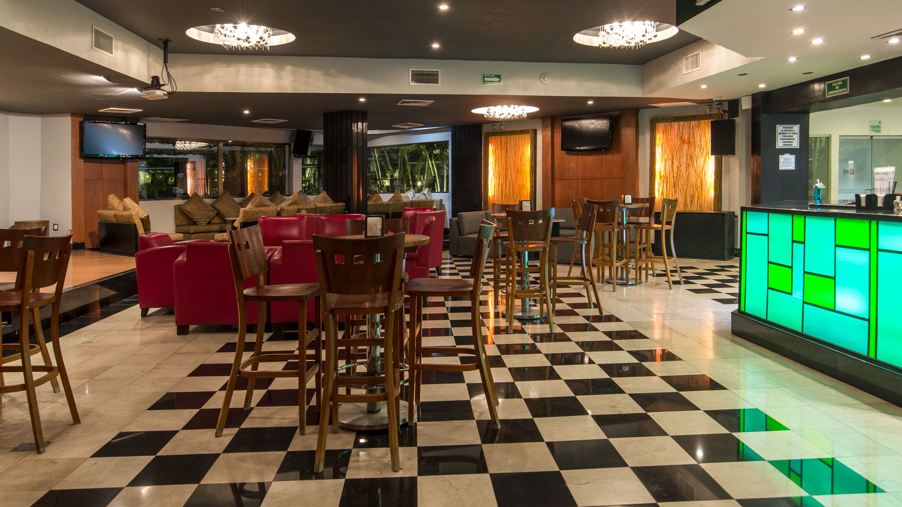 A hotel lobby with checkered flooring, red and wooden seating, a bar area with a green illuminated counter, and a large flat-screen TV mounted on the wall.