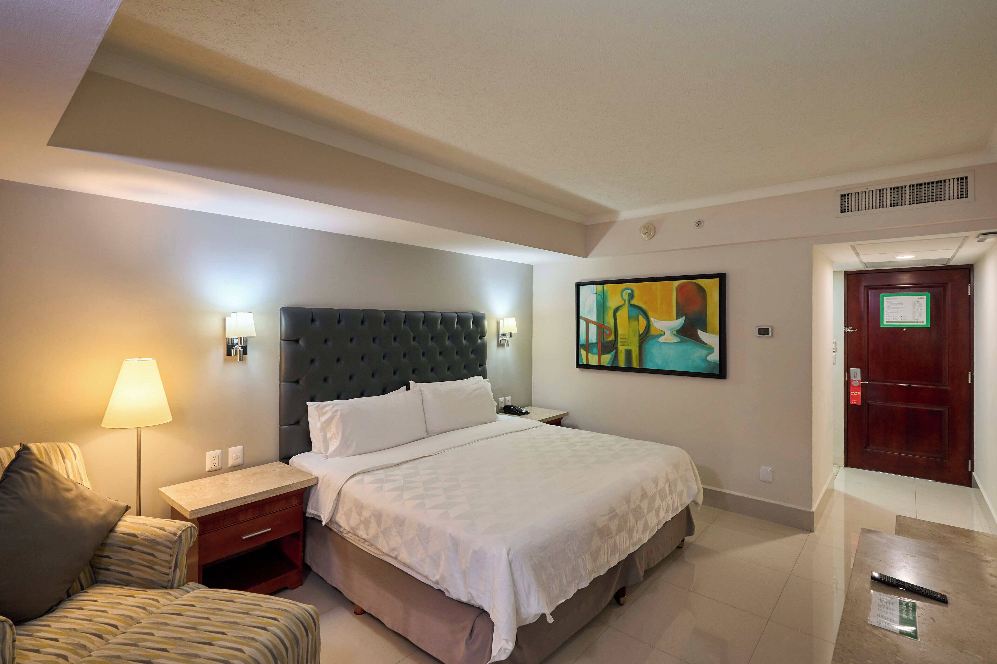 A hotel room with a large bed, a bedside table, a lamp, a chair, and a painting on the wall.