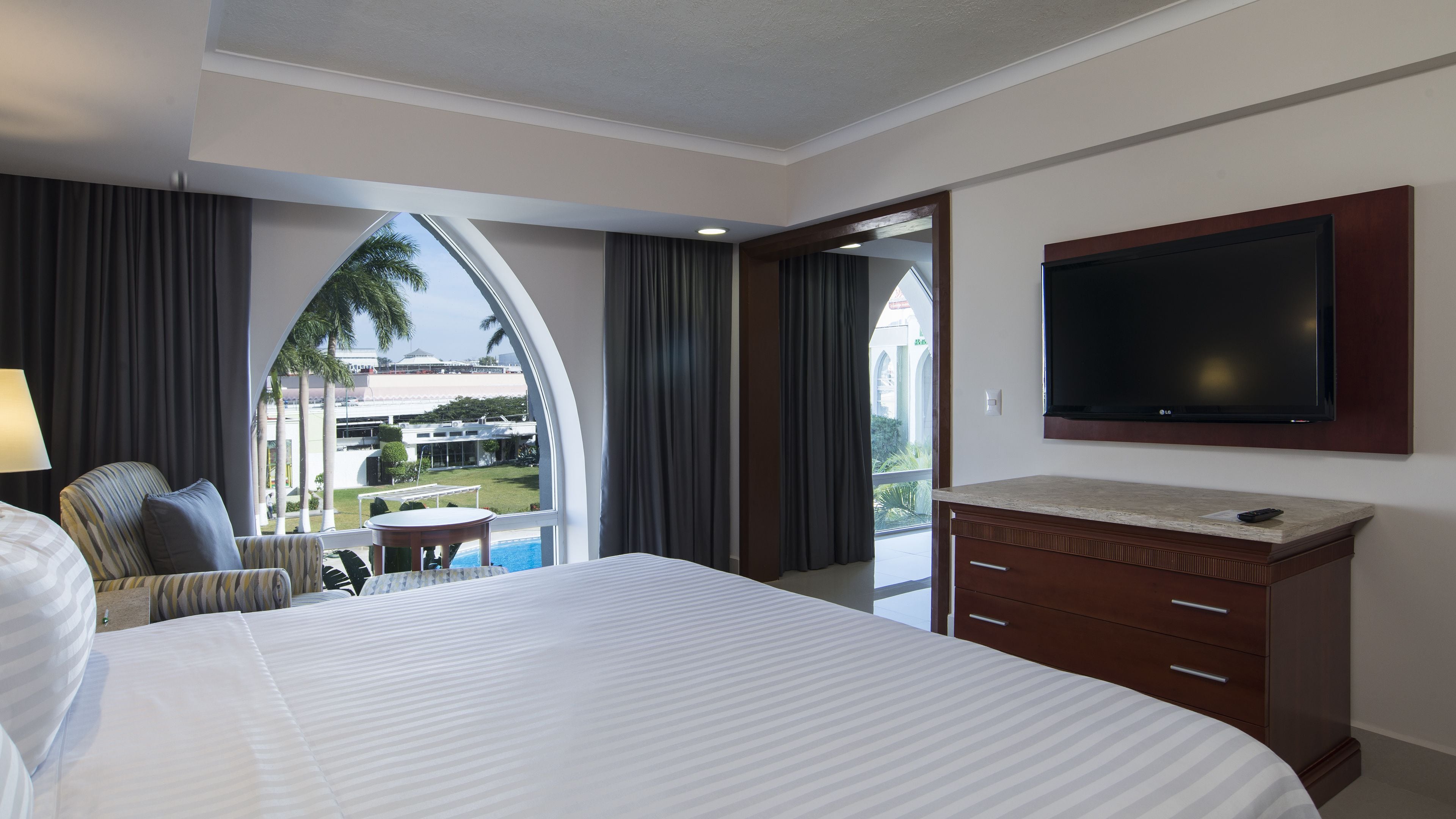 Une chambre d’hôtel avec un grand lit, un téléviseur fixé au mur, un bureau avec une chaise et une vue sur une piscine et des palmiers.