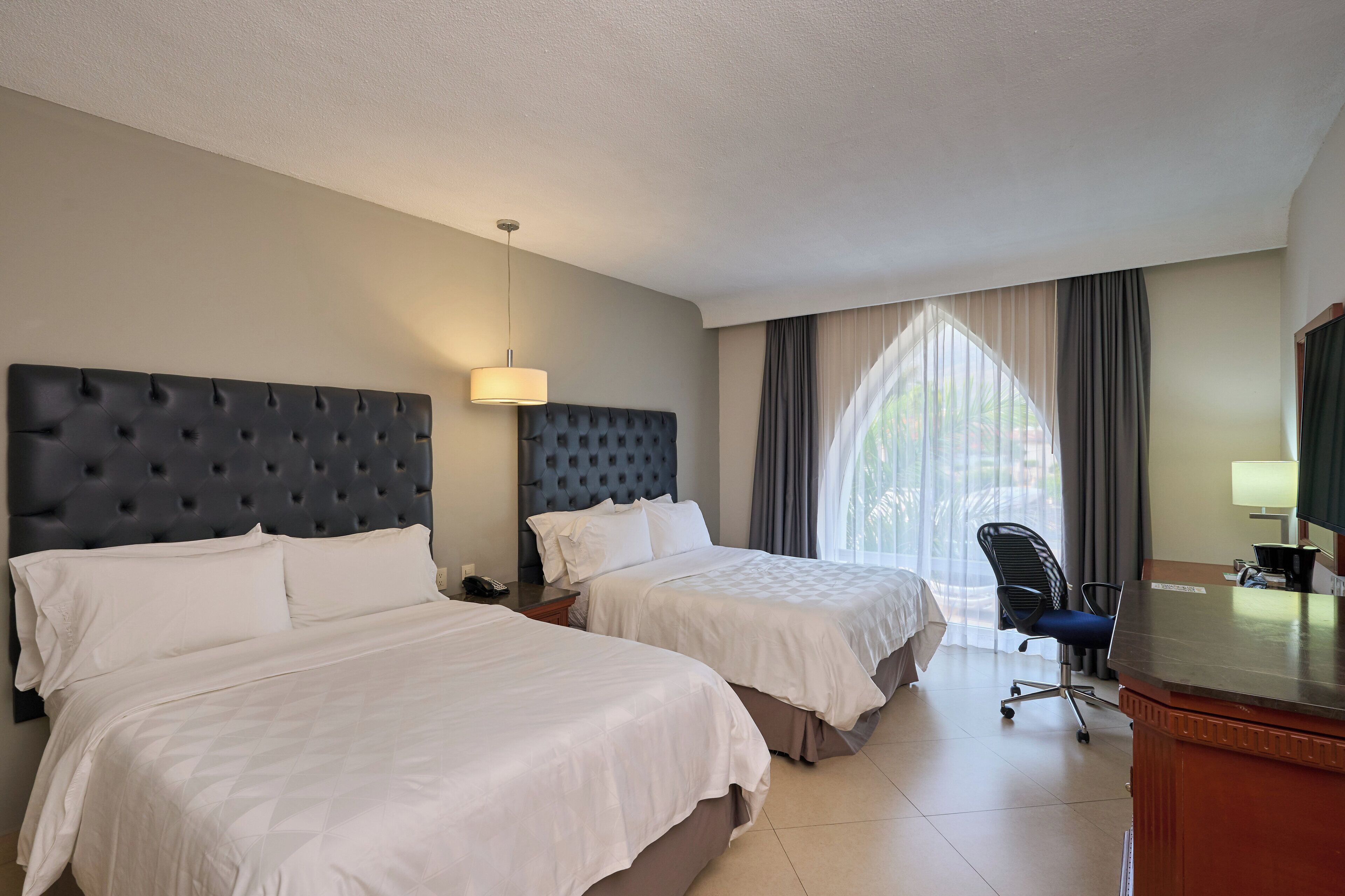 Habitación de hotel con dos camas, un escritorio, una silla y un ventanal con cortinas.