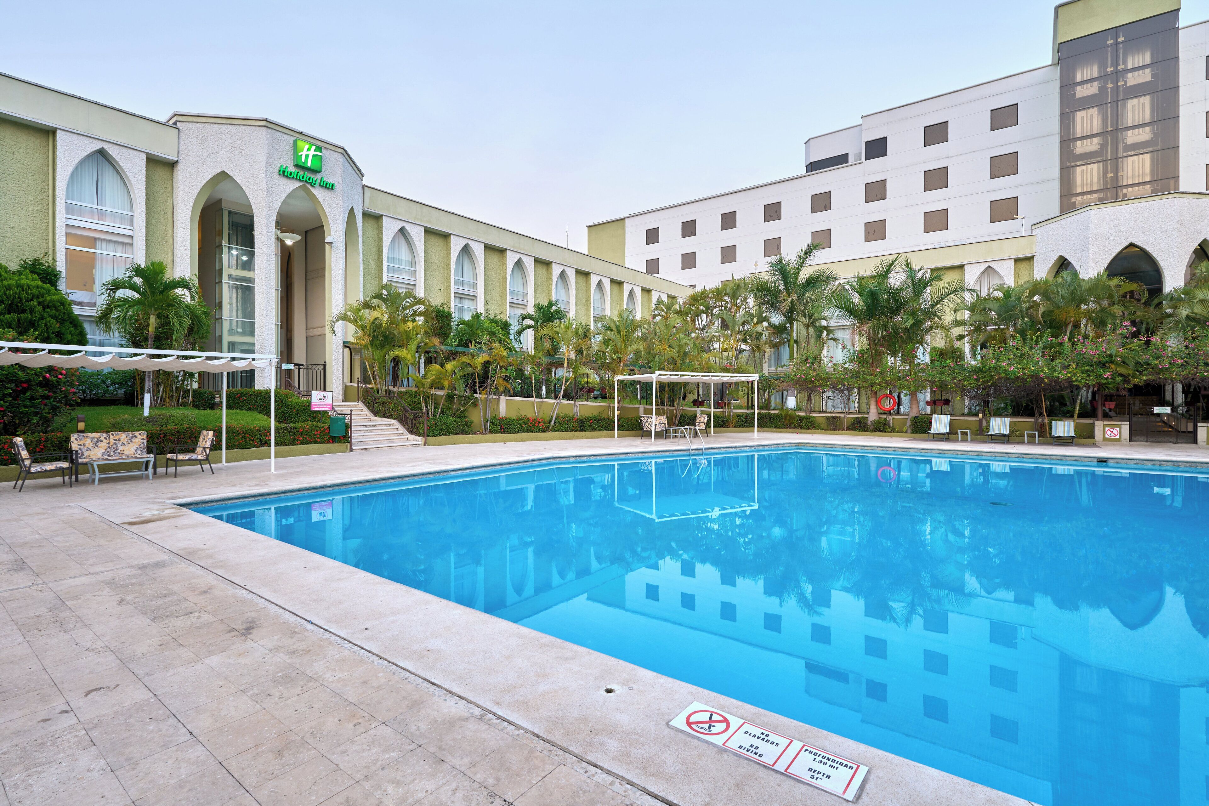 A swimming pool with lounge chairs and umbrellas, surrounded by palm trees and a hotel building with arched windows.
