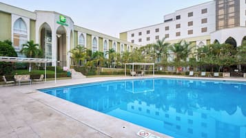 A swimming pool with lounge chairs and umbrellas, surrounded by palm trees and a hotel building with arched windows.