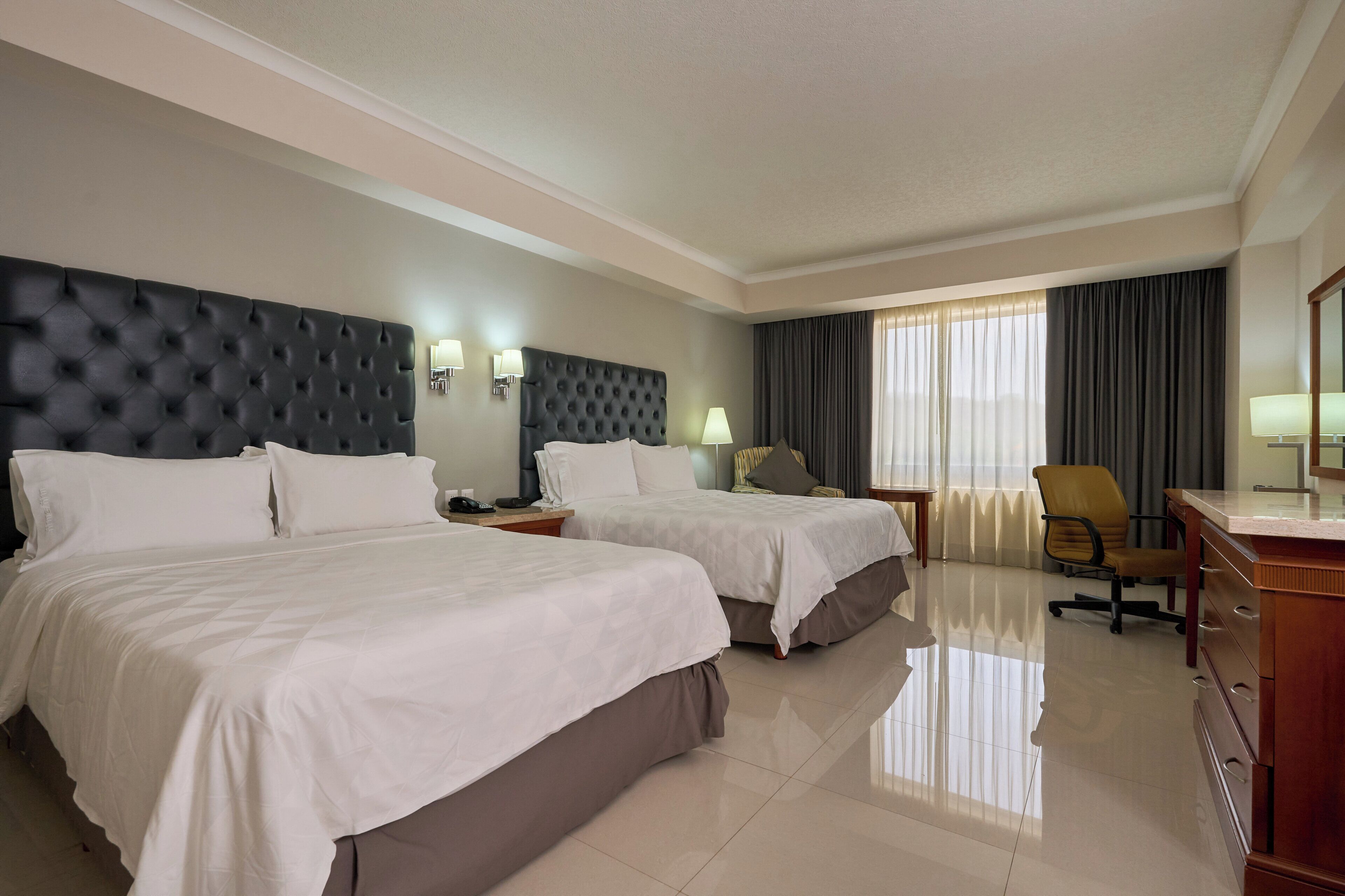A hotel room with two beds, a desk, a chair, and a large window with curtains.