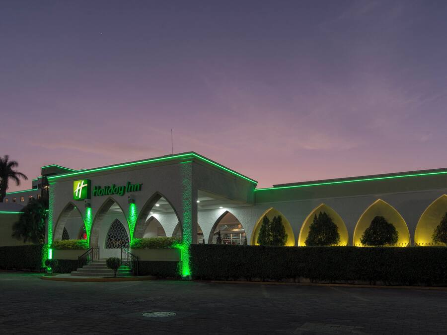 Un hotel Holiday Inn con fachada moderna, ventanas arqueadas y una palmera en primer plano.