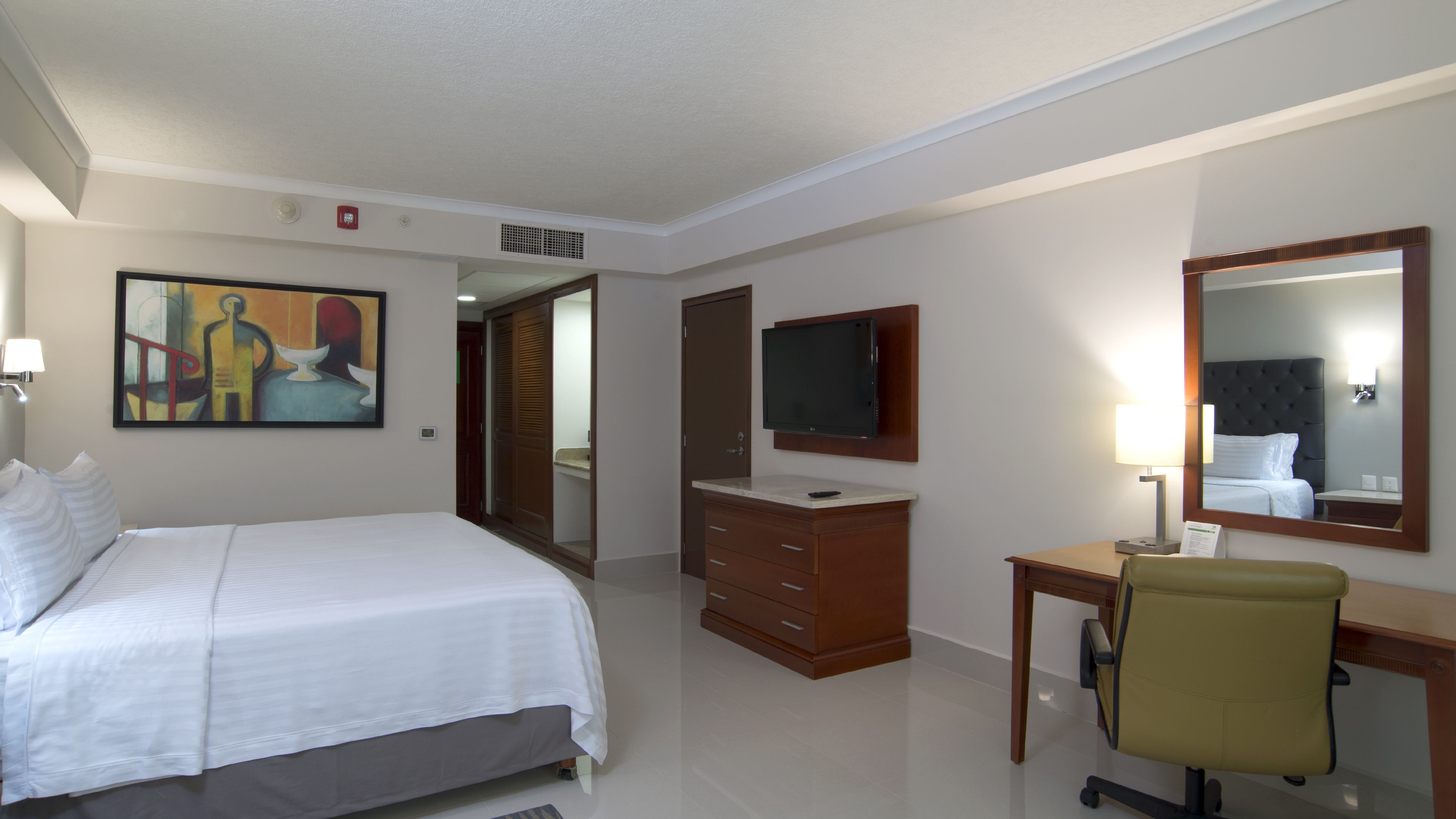 A hotel room with a bed, a desk with a chair, a television, a mirror, and a painting on the wall.