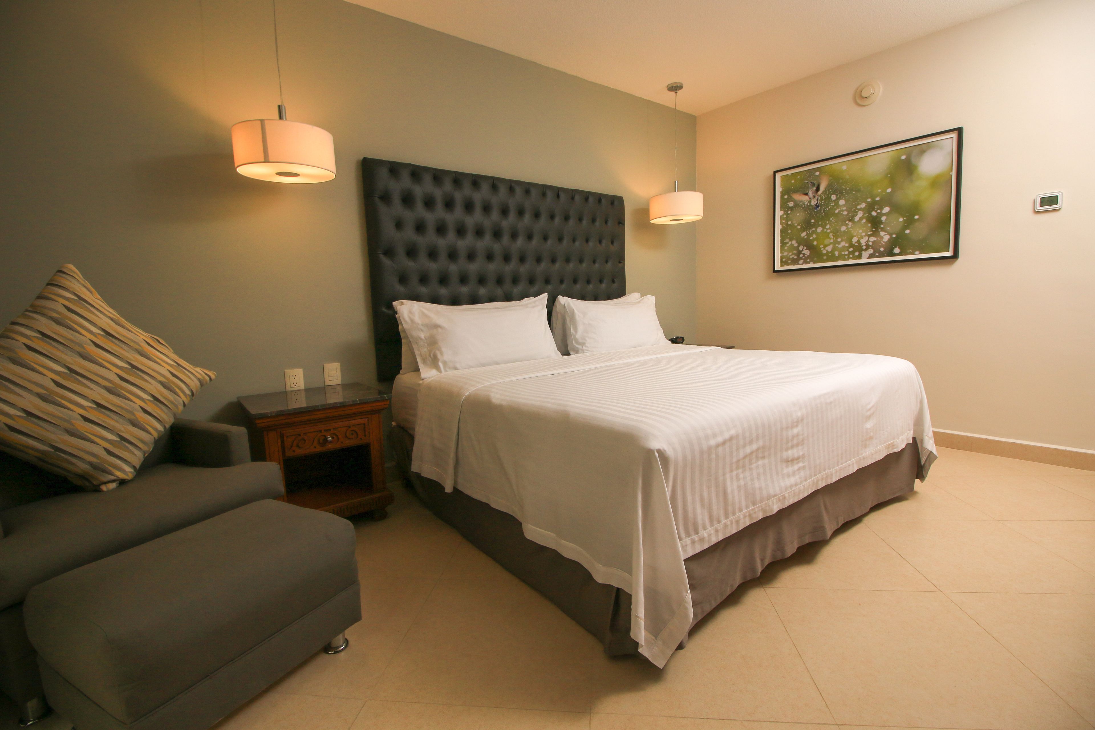 A hotel room with a bed, a nightstand, a lamp, a sofa, and a painting on the wall.