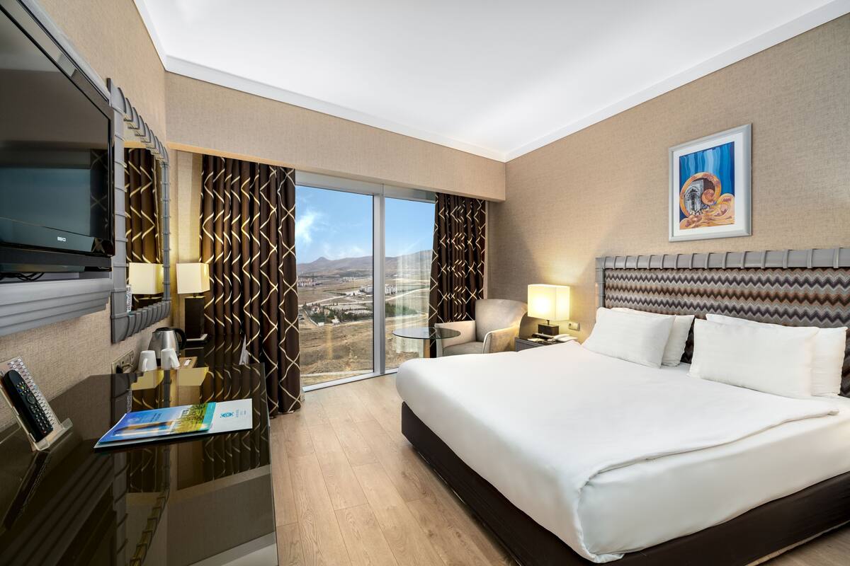 Ein Hotelzimmer mit einem großen Bett, einem Schreibtisch mit Computer, einem Fernseher und Blick auf die Stadt durch das Fenster.