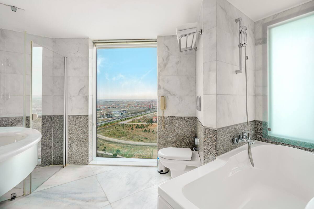 Ein modernes Badezimmer mit einer großen Badewanne, einer Dusche und einer Toilette. Eine Glastür führt zu einem Balkon mit Blick auf die Stadt.