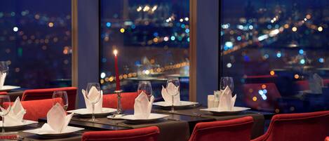 Un restaurante con mesas dispuestas para comer, sillas tapizadas en rojo y vistas a la ciudad a través de amplias ventanas.