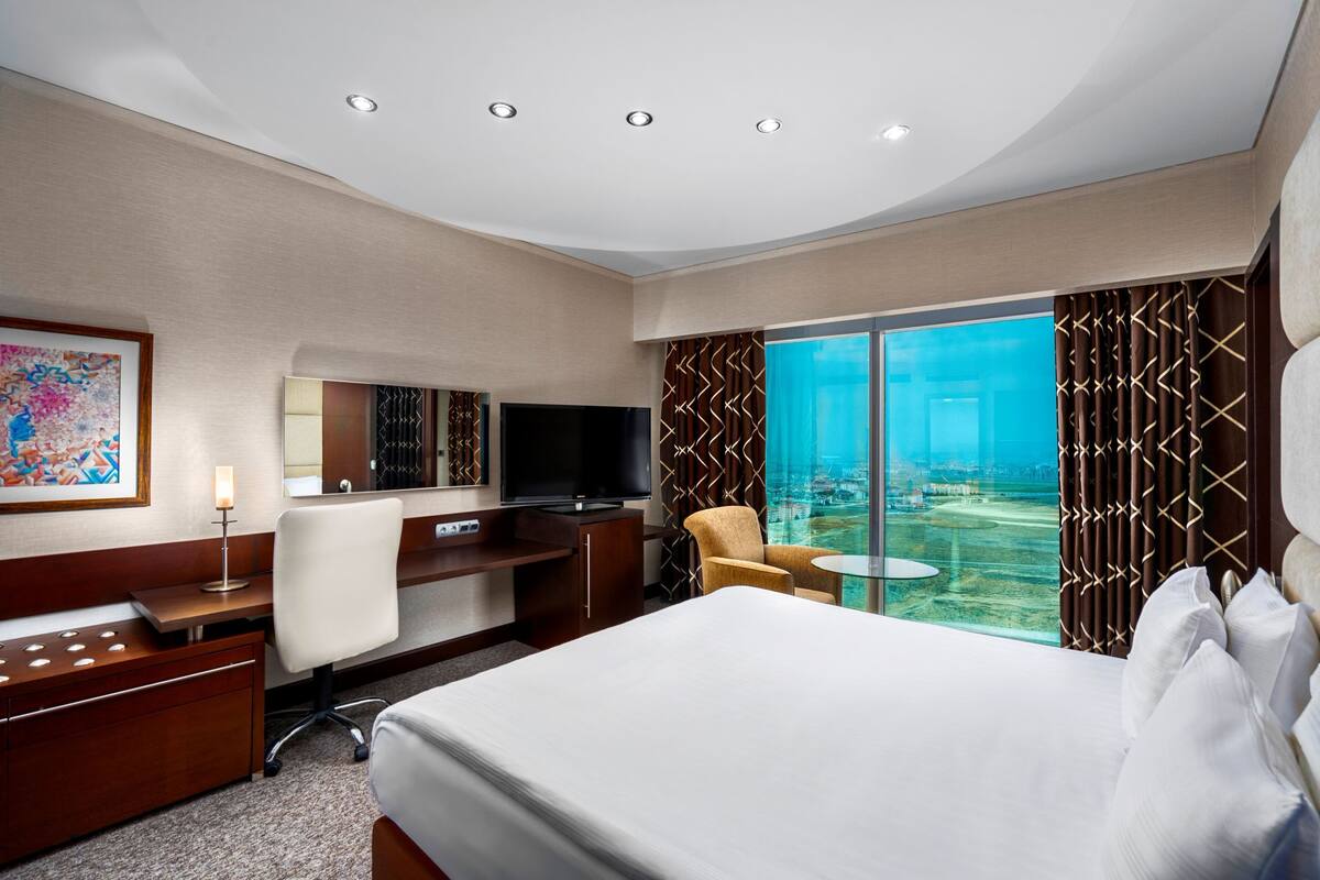 Ein Hotelzimmer mit Bett, Schreibtisch, Fernseher und einem großen Fenster mit Blick auf die Stadt.