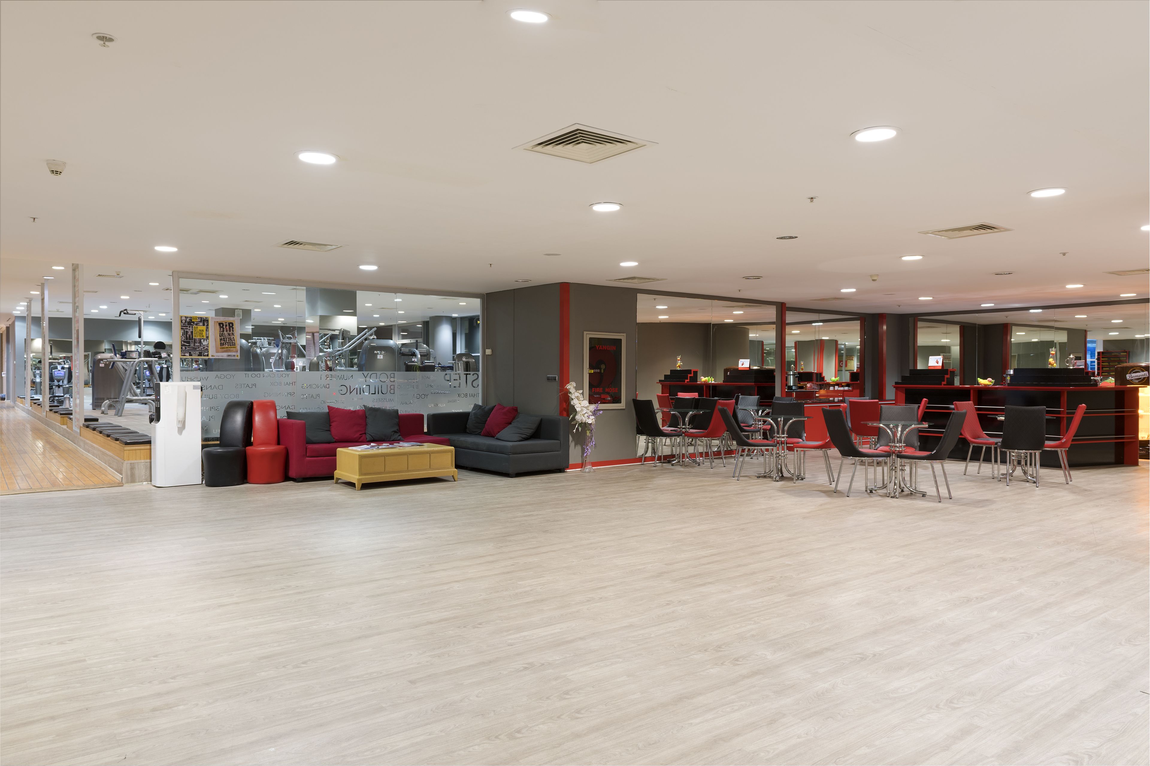Un gimnasio moderno con equipamiento cardiovascular, una zona de recepción y un área de descanso con sillas rojas y un sofá gris.