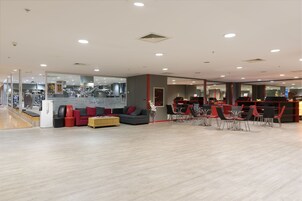Un gimnasio moderno con equipamiento cardiovascular, una zona de recepción y un área de descanso con sillas rojas y un sofá gris.