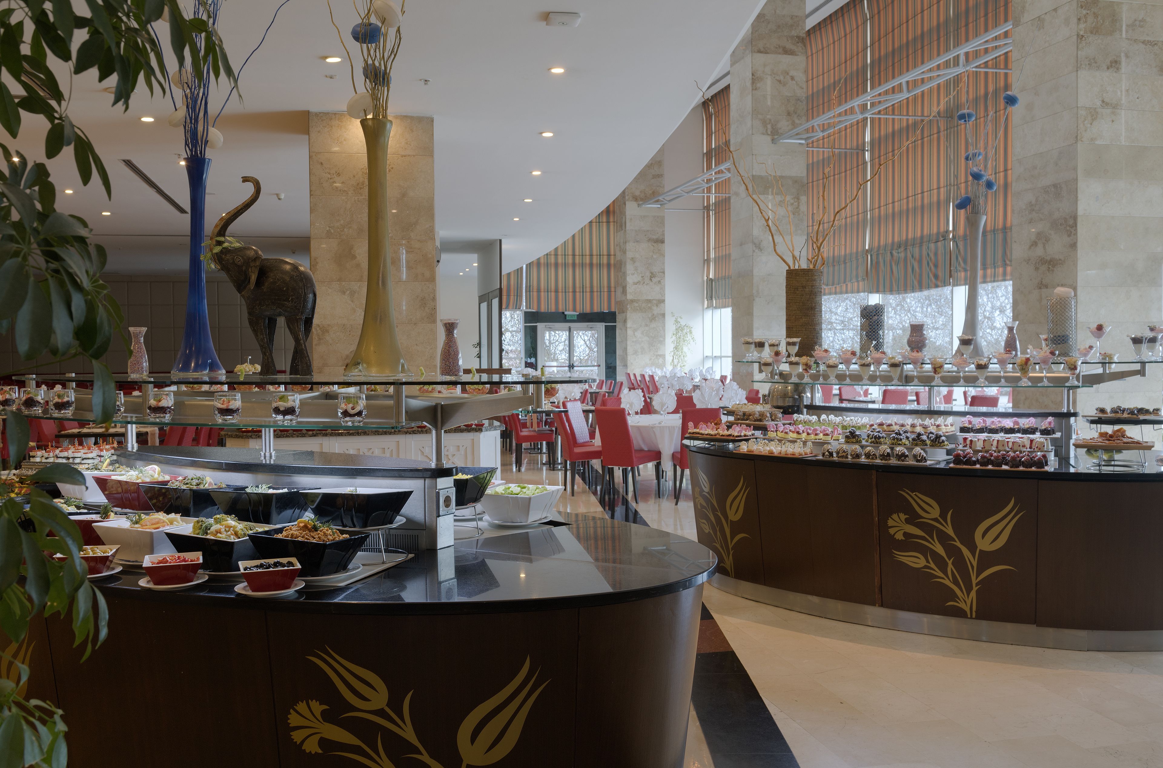 A buffet area with various food stations, a central decorative elephant sculpture, and a modern interior design with large windows and a curved ceiling.