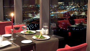 Uma mesa de jantar posta com pratos, copos e talheres, com uma vela e vista para o horizonte urbano à noite.