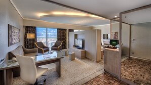 Quarto de hotel moderno com secretária de vidro, duas poltronas, televisão e minibar.