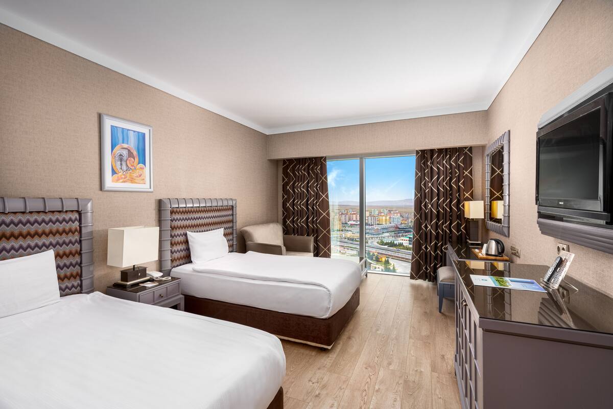 Ein Hotelzimmer mit zwei Betten, einem Schreibtisch, einem Fernseher und Blick auf die Stadt.