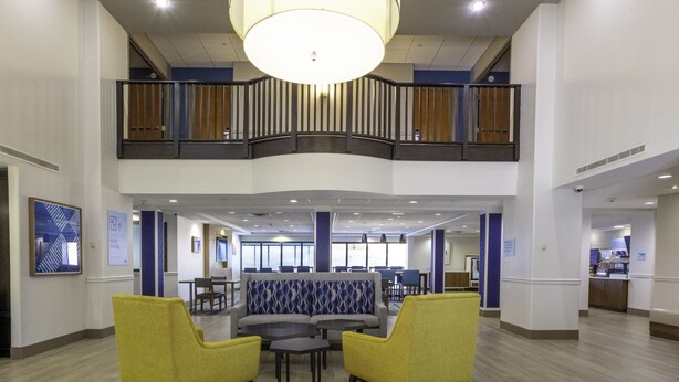Eine geräumige Lobby mit hoher Decke, einem zentralen Sitzbereich mit Sofas und Sesseln, einer Treppe zu einer oberen Ebene und einem Empfangstresen im Hintergrund.