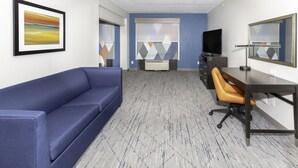 Ein Hotelzimmer mit einem blauen Sofa, einem Schreibtisch mit Stuhl, einem Flachbildfernseher und einem großen Fenster mit Jalousien.
