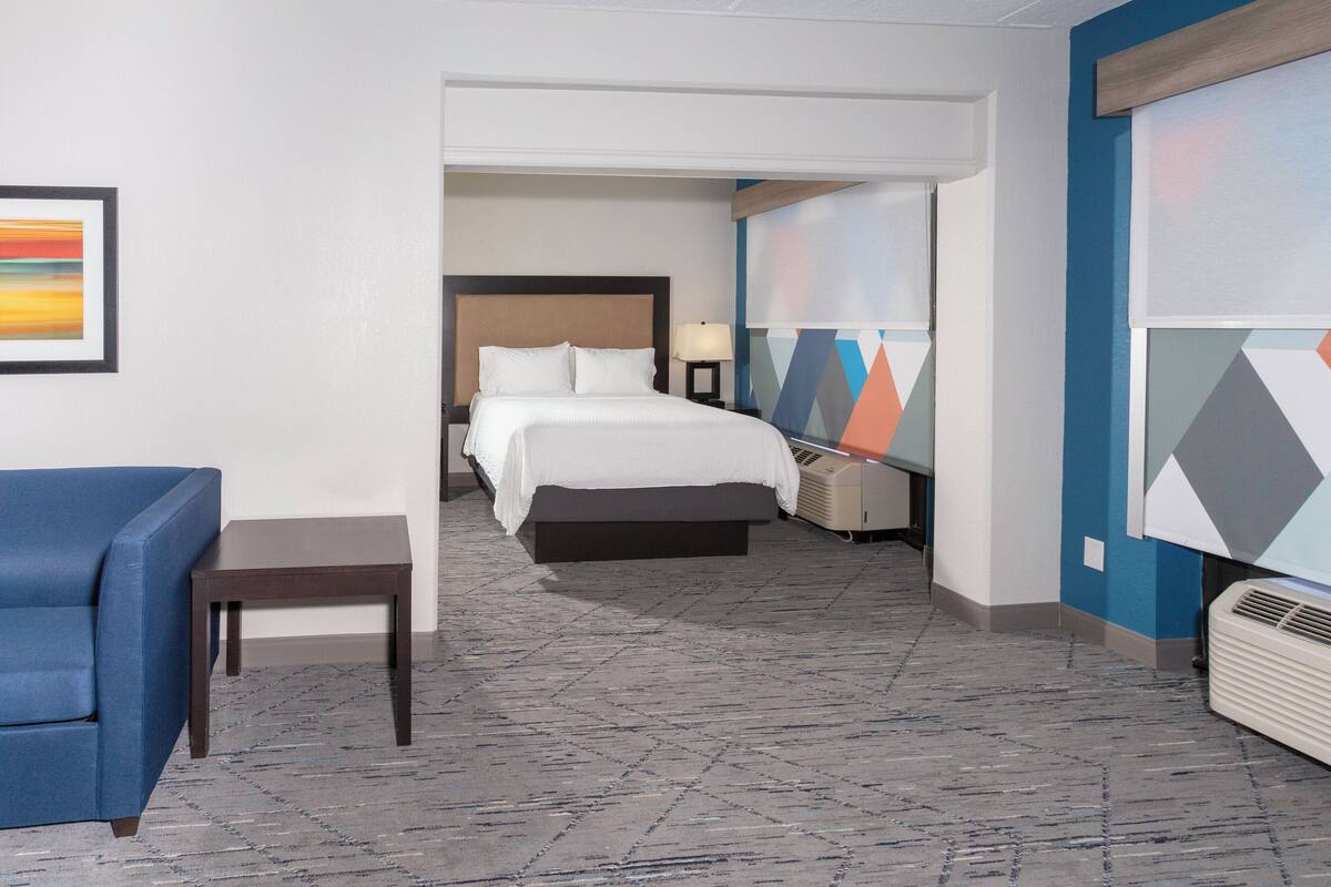 Une chambre d’hôtel avec un lit, une table de chevet, une lampe, un fauteuil bleu et un tapis à motifs géométriques.